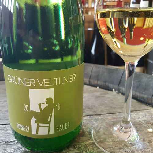 340316_0001 Grüner Veltliner 1 Liter von Norbert Bauer in grüner Flasche mit olivgrünem Etikett und Glas Weißwein daneben auf einem Holztisch